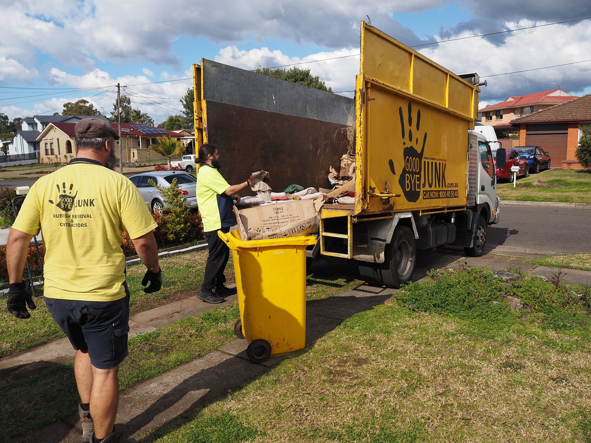 Two workers of Good Bye Junk Rubbish Removal loading cardboard boxes and other items from a yellow wheeled bin into the back the truck.  The scene is taking place on a residential street with houses, power lines, and parked cars visible in the background.