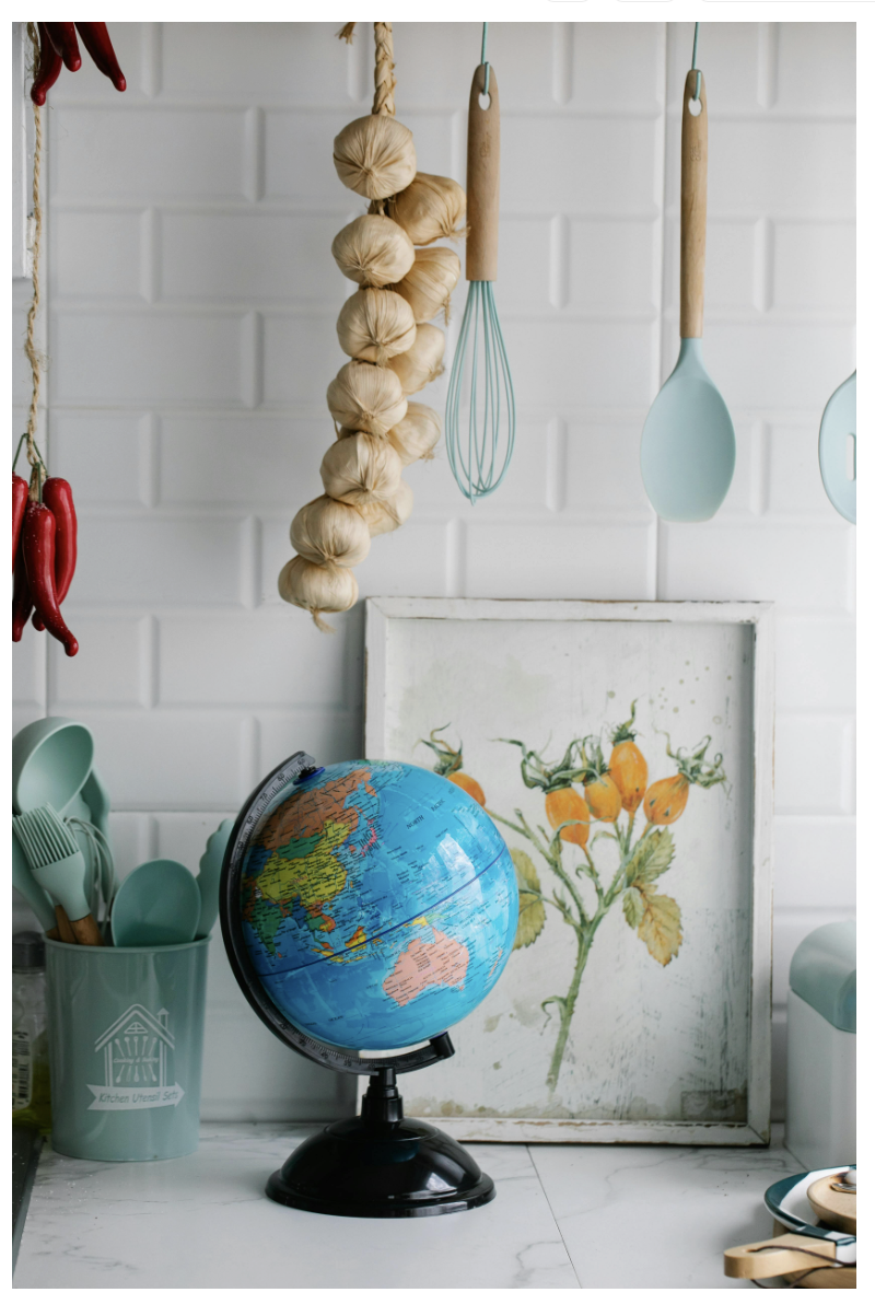 Kitchen counter with globe, botanical art, herbs and spices, and pastel utensils against a white tile backsplash.