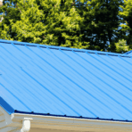 Blue metal roof with diagonal ridges, adjacent to a brick chimney. Evergreen trees in the background, conveying a bright, clear day.