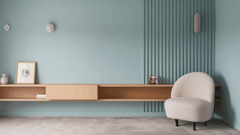 minimalist living room with blue paneled wall, floating wood shelf, decor, and cream accent chair