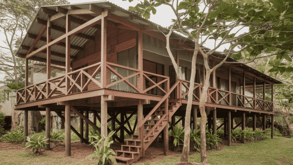 raised wooden house on stilts with wraparound porch and exterior staircase surrounded by trees